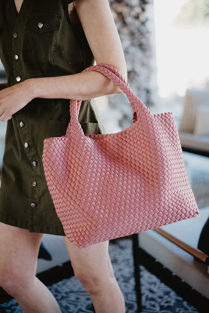 A person holds the Katydid Blush Woven Tote Bag by its handles while wearing a sleeveless, button-up olive green dress. The softly blurred background features indoor decor.