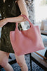 A person holds the Katydid Blush Woven Tote Bag by its handles while wearing a sleeveless, button-up olive green dress. The softly blurred background features indoor decor.