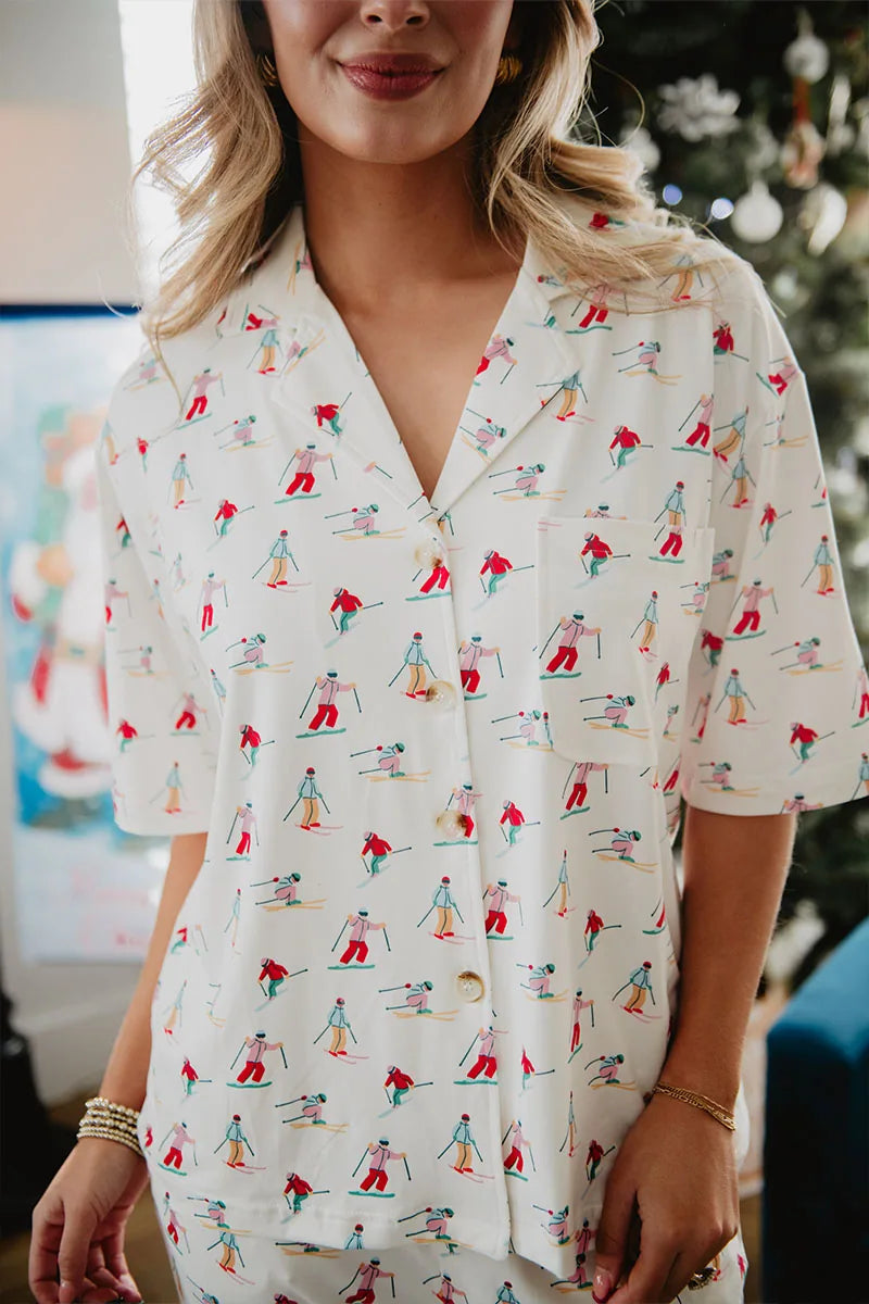A woman wears the Katydid Apres Ski Matching Pajama Pants Set, a short-sleeved, white button-up featuring playful skiers in red and blue. A blurred decorated Christmas tree appears in the background.