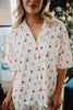 A woman wears the Katydid Apres Ski Matching Pajama Pants Set, a short-sleeved, white button-up featuring playful skiers in red and blue. A blurred decorated Christmas tree appears in the background.