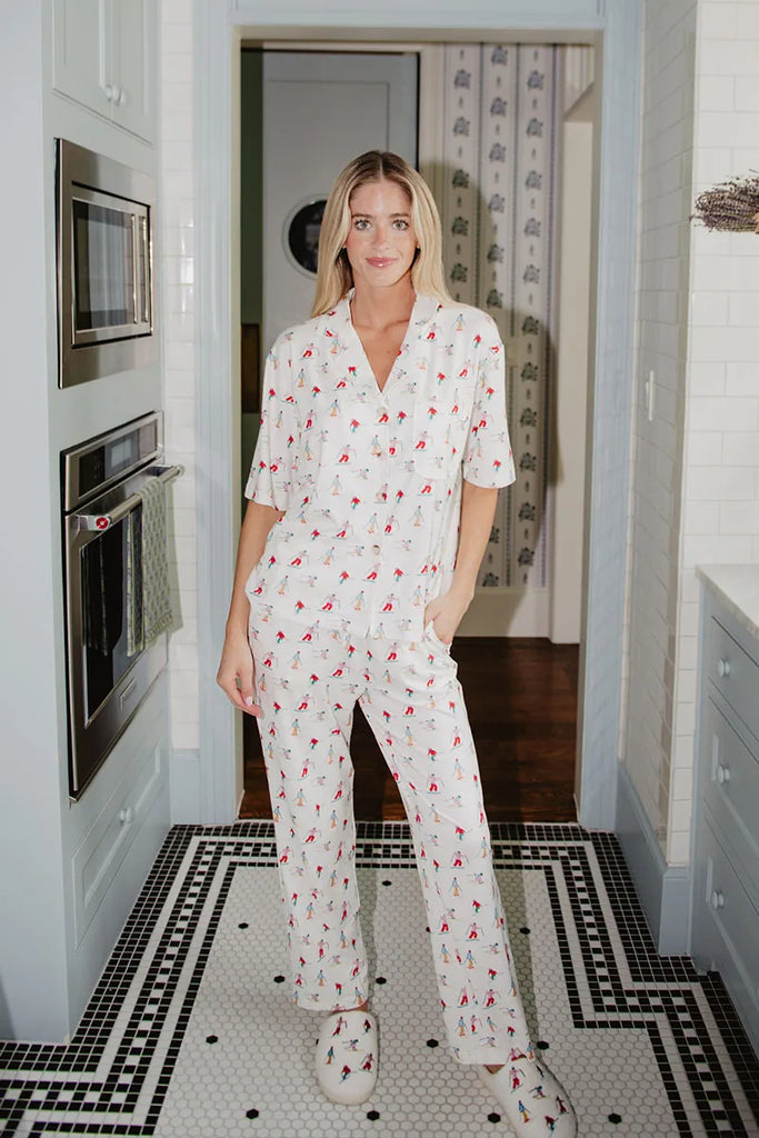 A woman smiles in a kitchen with white cabinets and patterned tile floor, wearing the Katydid Apres Ski Matching Pajama Pants Set—red floral print with matching white slippers—one hand tucked in her pocket.