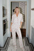 A woman smiles in a kitchen with white cabinets and patterned tile floor, wearing the Katydid Apres Ski Matching Pajama Pants Set—red floral print with matching white slippers—one hand tucked in her pocket.