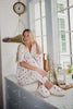 A woman wearing the Katydid Apres Ski Matching Pajama Pants Set and slippers sits on a kitchen counter by a window, smiling. Drinks, snacks, and kitchen items are nearby. A clock hangs on the tiled wall above her.