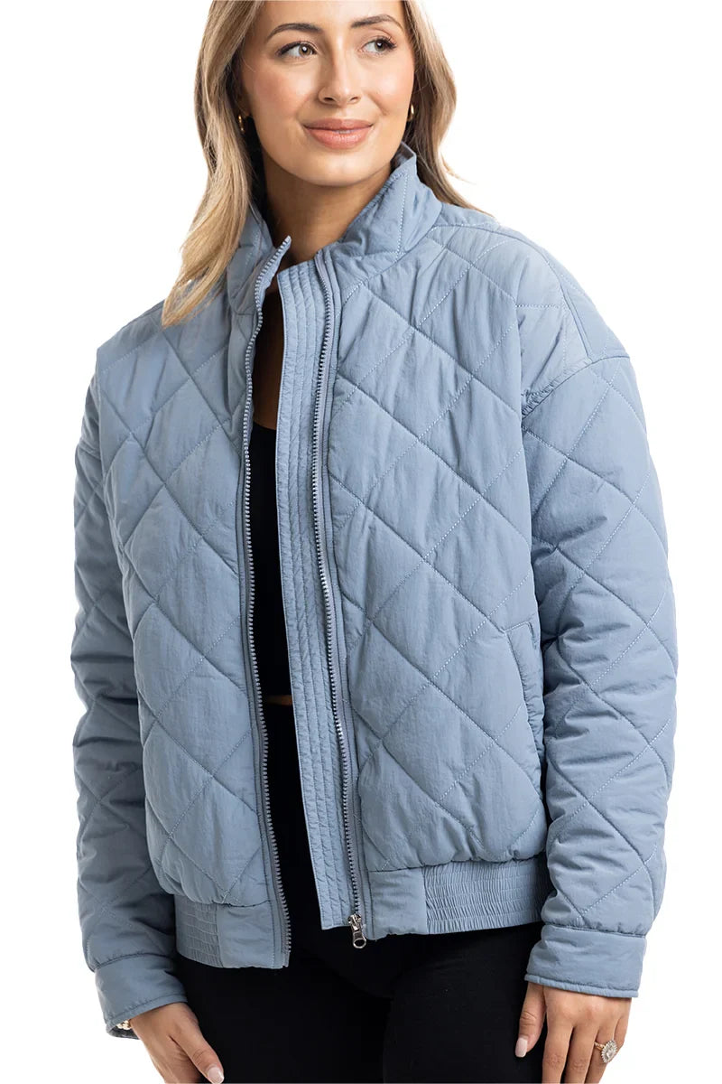 A woman models the Katydid Steel Blue Sporty Diamond Quilted Womens Jacket, unzipped over a black top, with loosely styled light hair, standing against a plain white background.