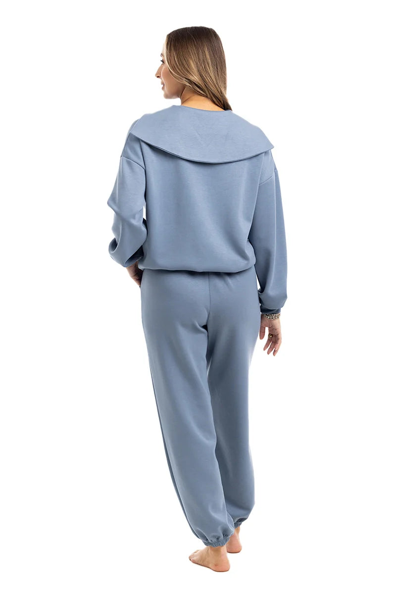 A woman faces away, barefoot on a white background, wearing the Katydid Steel Blue Matching Sets Womens Pants—a loose, light blue set with a wide-collared sweatshirt and jogger pants for a relaxed, comfortable look.