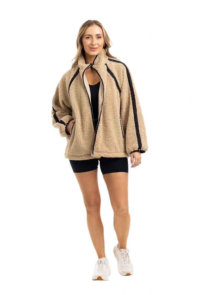 A woman with long, light hair smiles in a Katydid Tan Sherpa Lace Trim Jacket, paired with black shorts and white sneakers, posing against a plain white background.