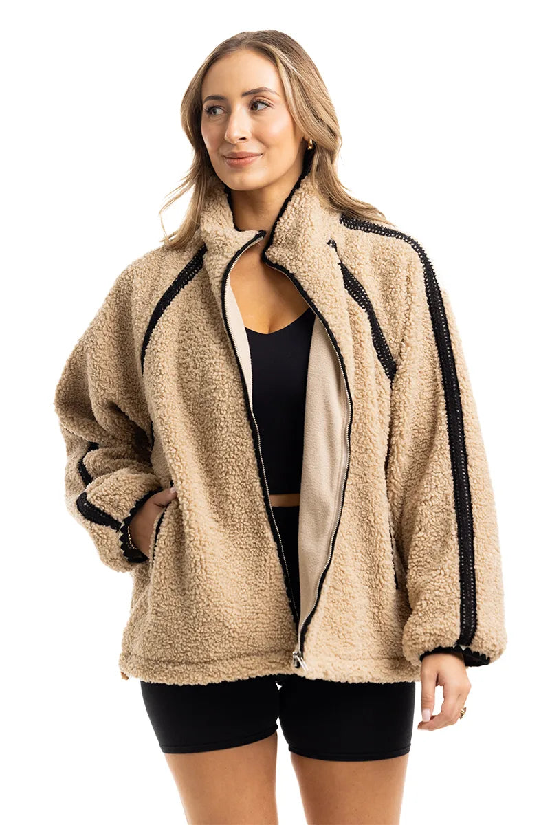A woman models the Katydid Tan Sherpa Lace Trim Jacket over a black crop top and shorts, standing with one hand in her pocket against a white background to showcase this cozy fall piece.