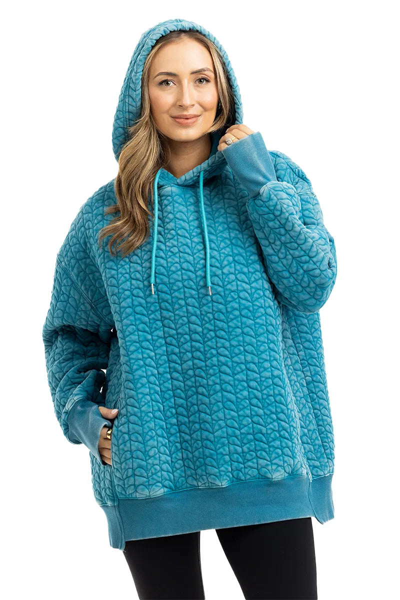 A woman smiles, wearing the Katydid Deep Teal Heart Quilted Garment Washed Hoodie with thumbholes, paired with black pants for a casual-chic style.