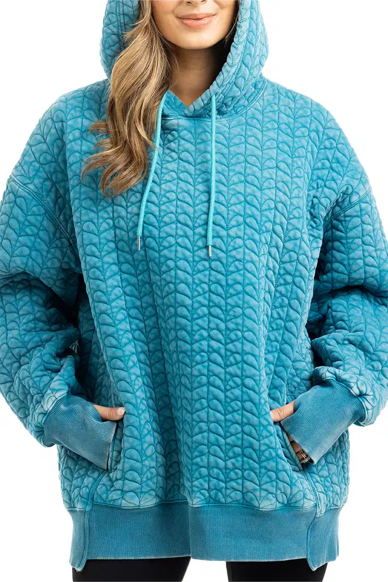 A person wears the Katydid Deep Teal Heart Quilted Garment Washed Hoodie with a drawstring hood and front pocket, their hands tucked in, and only part of their face visible.