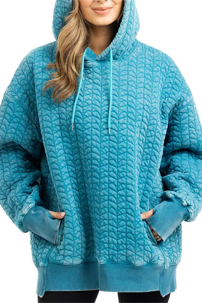 A person wears the Katydid Deep Teal Heart Quilted Garment Washed Hoodie with a drawstring hood and front pocket, their hands tucked in, and only part of their face visible.