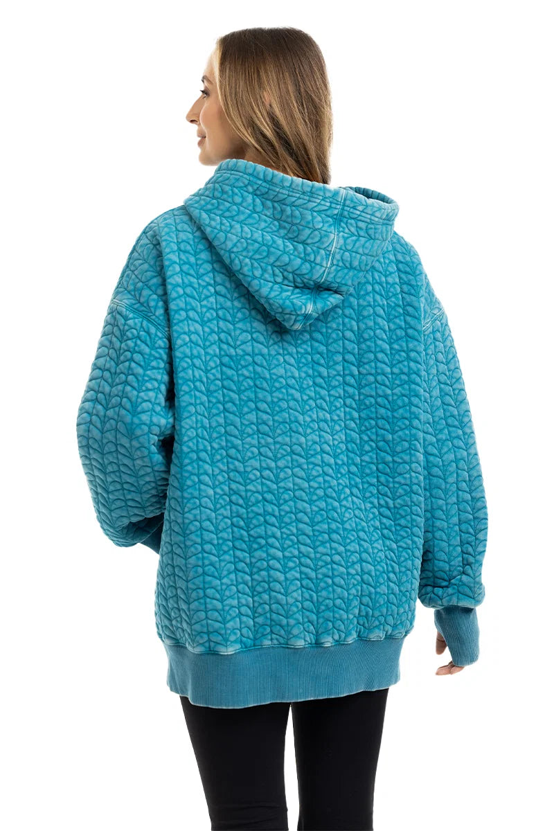 A woman with long hair is wearing the Katydid Deep Teal Heart Quilted Garment Washed Hoodie, facing away to show its back. She pairs the hoodie with black pants.