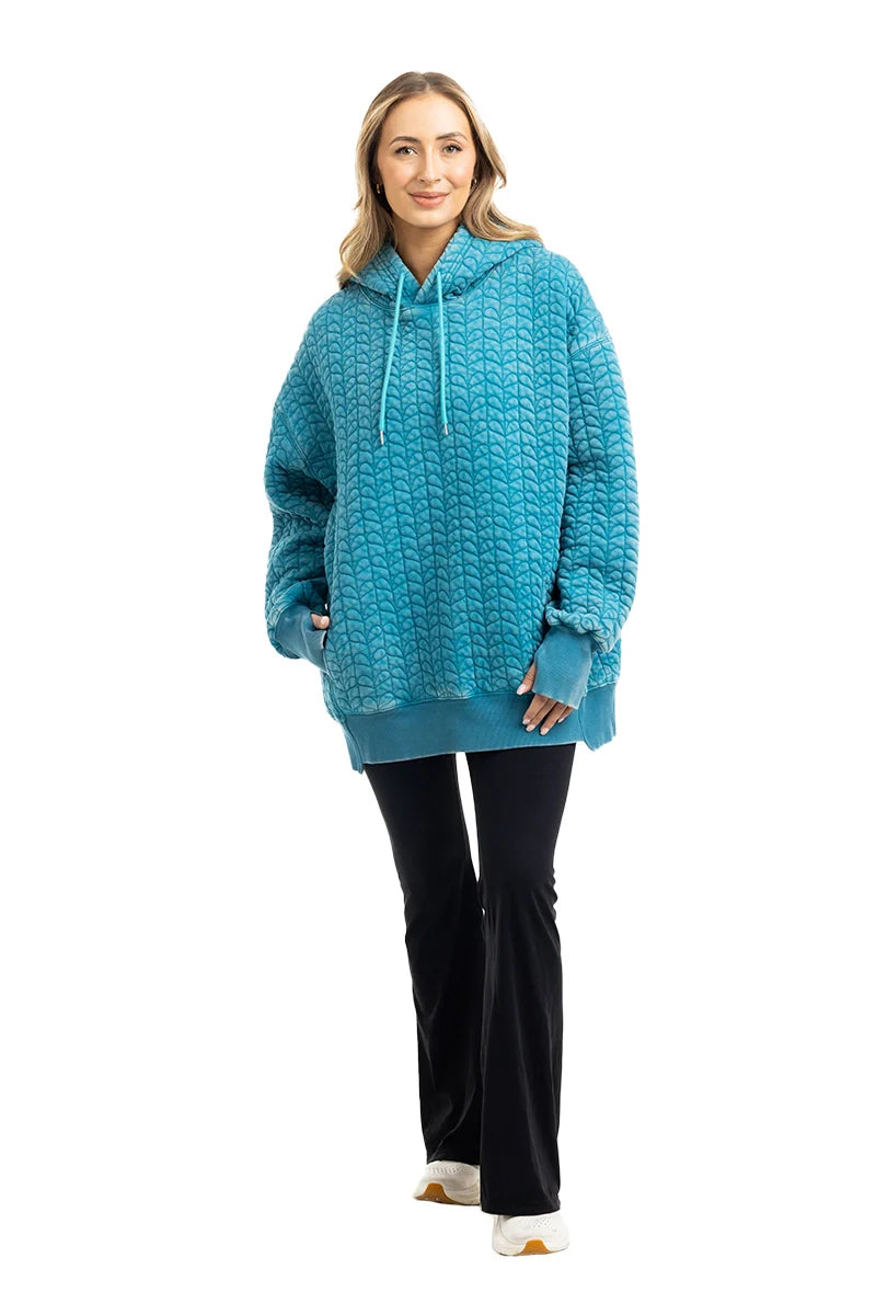 A woman smiles against a plain white background, wearing the Katydid Deep Teal Heart Quilted Garment Washed Hoodie with a relaxed fit, paired with black flared pants and white shoes.