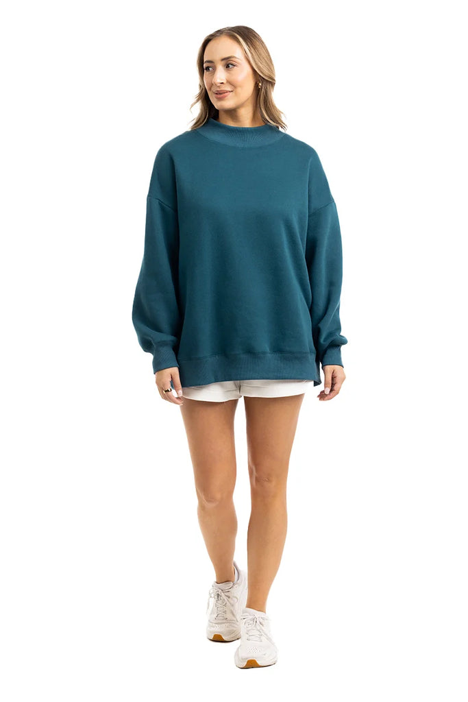 A woman with light brown hair smiles softly while looking off to the side, wearing the Katydid Deep Teal Essential Mockneck Sweatshirt, light shorts, and white sneakers against a white background—capturing effortless fall style.
