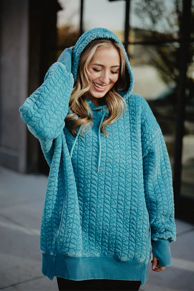 A woman with long blonde hair smiles outdoors, touching her hood while wearing the Katydid Deep Teal Heart Quilted Garment Washed Hoodie. The relaxed fit features a heart-quilted pattern, with a blurred building and windows in the background.