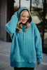 A woman with long blonde hair smiles outdoors, touching her hood while wearing the Katydid Deep Teal Heart Quilted Garment Washed Hoodie. The relaxed fit features a heart-quilted pattern, with a blurred building and windows in the background.
