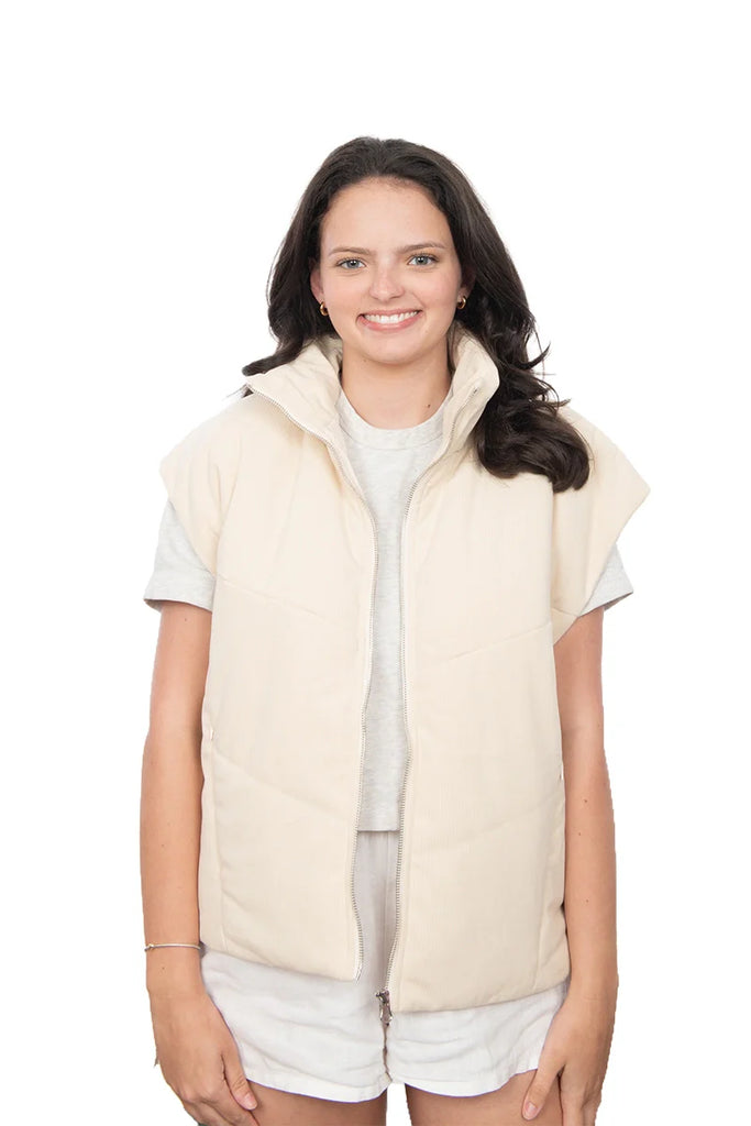 A woman with long brown hair smiles at the camera, wearing the Katydid Cloud White Corduroy Puffer Vest over a short-sleeve gray shirt and light shorts, standing against a plain white background.