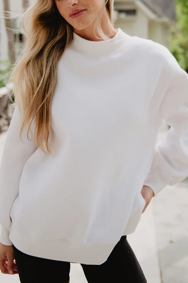 A woman with long blonde hair wears the Katydid Cloud White Essential Mockneck Sweatshirt and black pants, standing outdoors with one hand in her pocket.