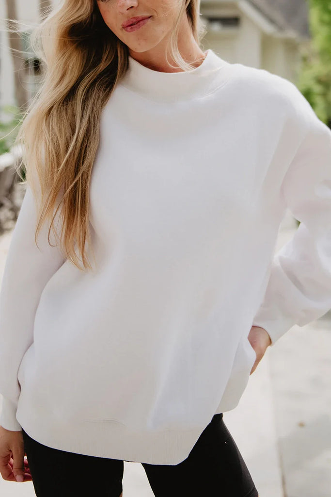 A woman with long blonde hair wears the Katydid Cloud White Essential Mockneck Sweatshirt and black pants, standing outdoors with one hand in her pocket.