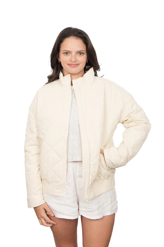 A young woman smiles with one hand in her pocket, wearing the Katydid Cloud White Sporty Diamond Quilted Jacket over a light shirt and shorts against a white background.