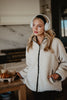 A woman in a Katydid Cream Womens Sherpa Lace Trim Jacket and white headphones stands thoughtfully in a kitchen. Iced coffee drinks and croissants in a takeout tray sit on the counter beside her.