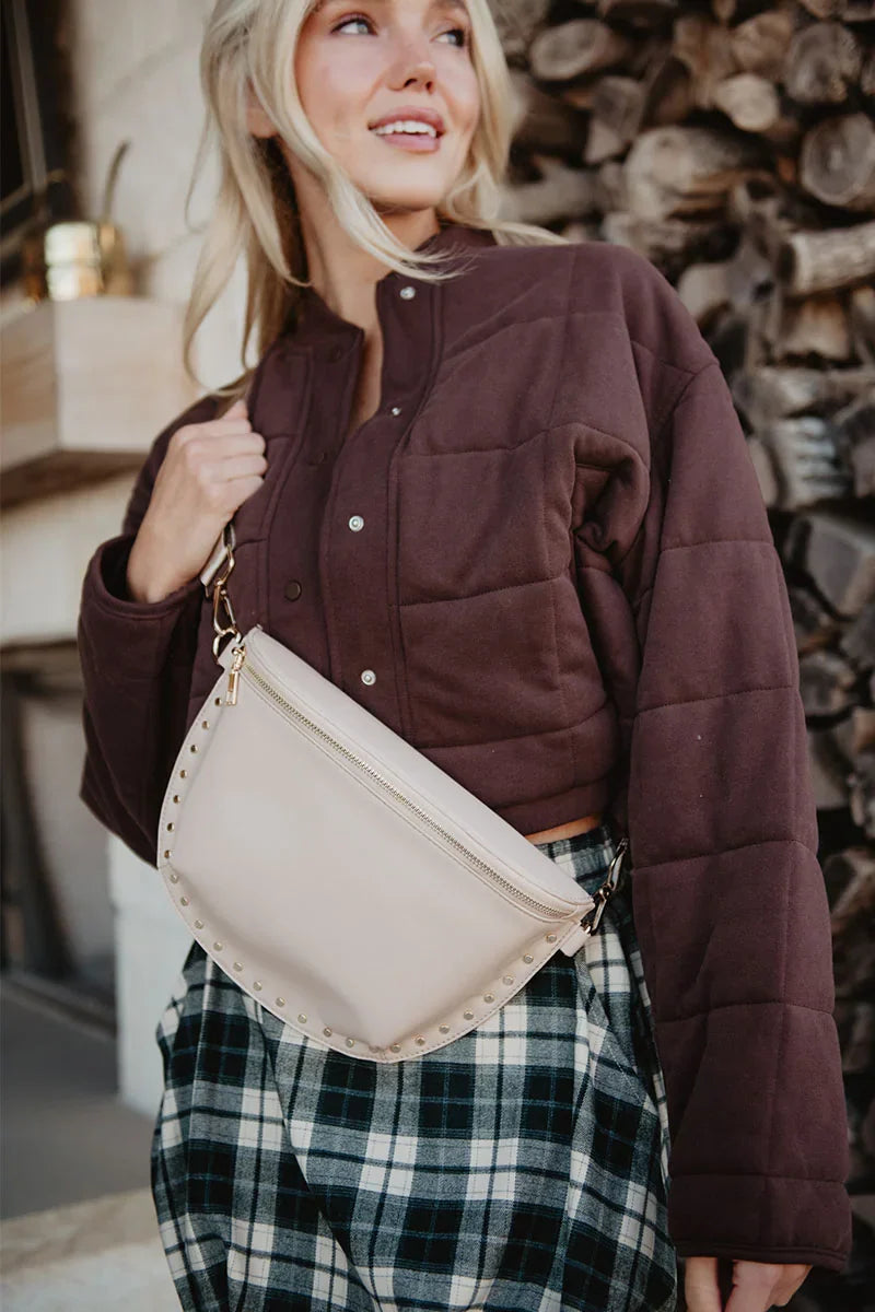 A woman smiles before stacked firewood in a cropped, quilted brown jacket and plaid skirt, carrying the Katydid Tan Gold-Studded Structured Crossbody—the perfect fall accessory.