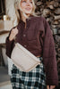 A woman smiles before stacked firewood in a cropped, quilted brown jacket and plaid skirt, carrying the Katydid Tan Gold-Studded Structured Crossbody—the perfect fall accessory.