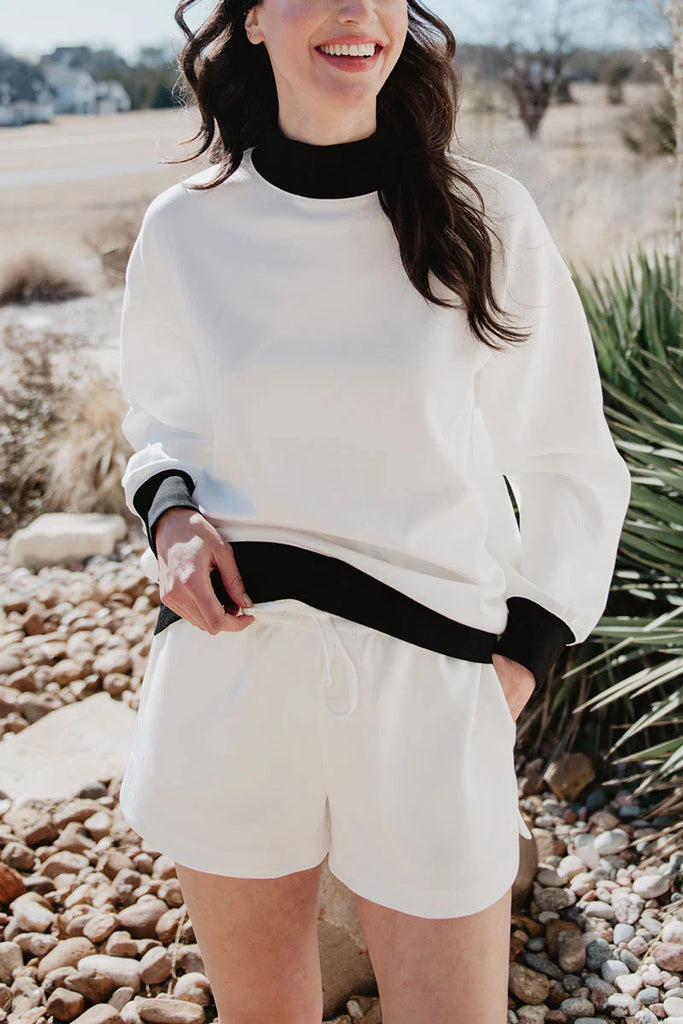 A woman smiles outdoors on a rocky path, wearing the Katydid White  Black Duo Mockneck Set—featuring a white sweatshirt with black collar and cuffs, and matching drawstring shorts. Shrubs and a road appear in the background.