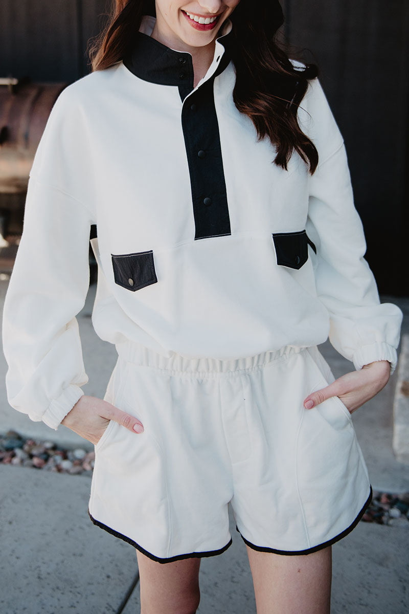 A woman smiles outdoors with her hands in her pockets, wearing the Katydid White  Black Quarter Snap Set—a sporty lounge set featuring a long-sleeved pullover with black buttons and pocket flaps, paired with matching shorts.