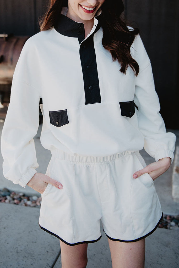 A woman smiles outdoors with her hands in her pockets, wearing the Katydid White  Black Quarter Snap Set—a sporty lounge set featuring a long-sleeved pullover with black buttons and pocket flaps, paired with matching shorts.
