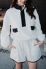 A woman smiles outdoors with her hands in her pockets, wearing the Katydid White  Black Quarter Snap Set—a sporty lounge set featuring a long-sleeved pullover with black buttons and pocket flaps, paired with matching shorts.