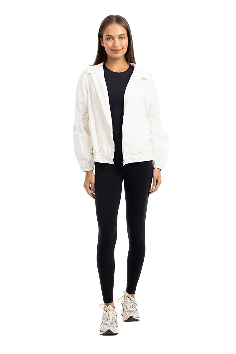 A woman faces forward against a white background, wearing the Katydid Cloud White Lightweight Hooded Jacket, paired with black leggings and white sneakers.