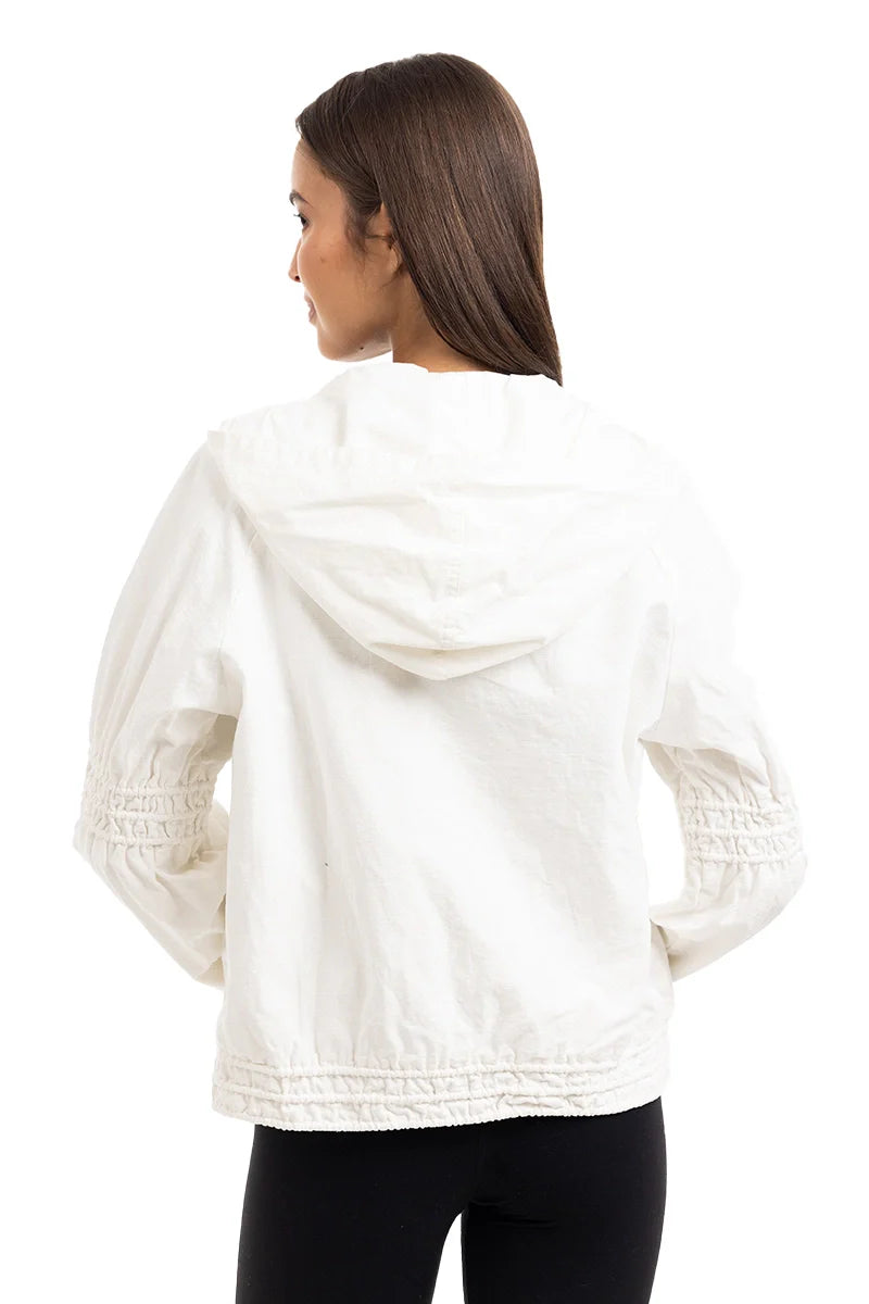 A woman with straight brown hair is seen from the back wearing the Katydid Cloud White Lightweight Hooded Jacket, featuring cinched sleeves and hem, paired with black pants against a plain white background.