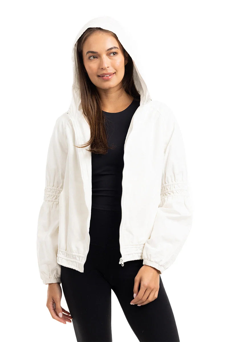 A woman models the Katydid Cloud White Lightweight Hooded Jacket over a black top and pants, standing against a white background and smiling slightly while looking to the side.
