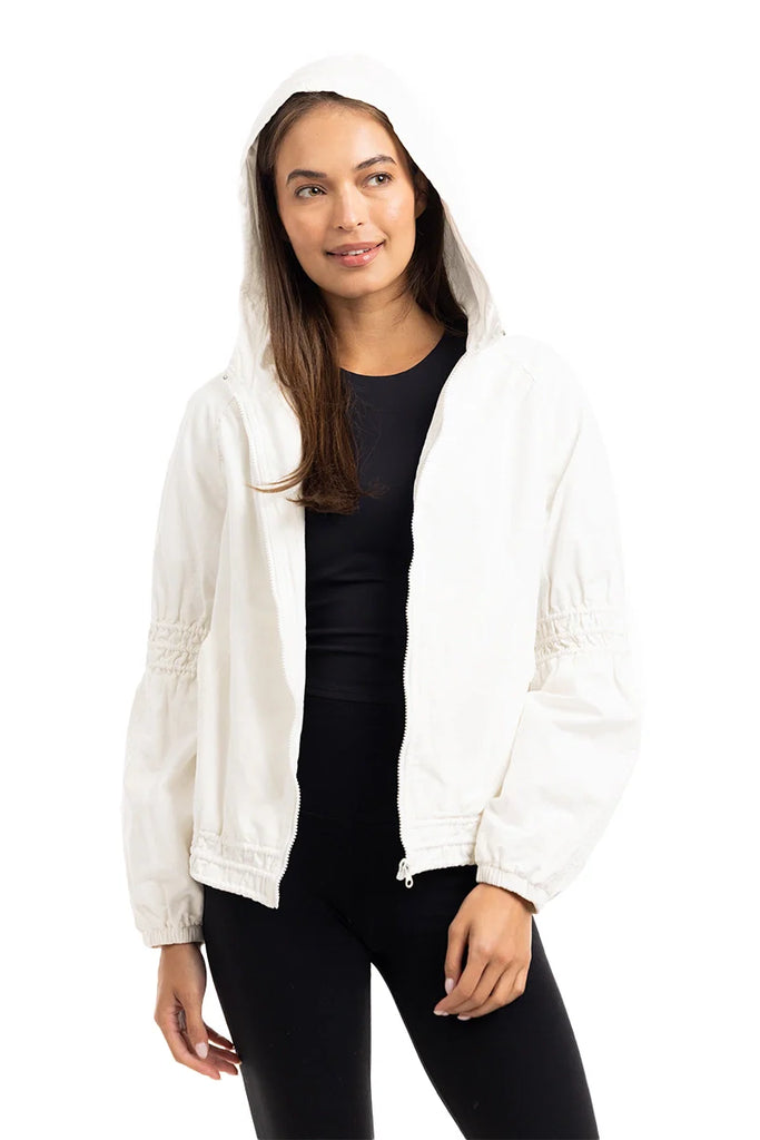A woman models the Katydid Cloud White Lightweight Hooded Jacket over a black top and pants, standing against a white background and smiling slightly while looking to the side.