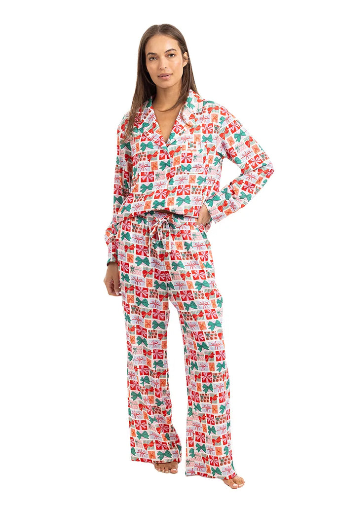 A woman wears the Katydid Wrapped Presents with Bows Pajama Pants Set, featuring a long-sleeve button-up top and drawstring pants adorned with colorful red, green, and pink gift box patterns.