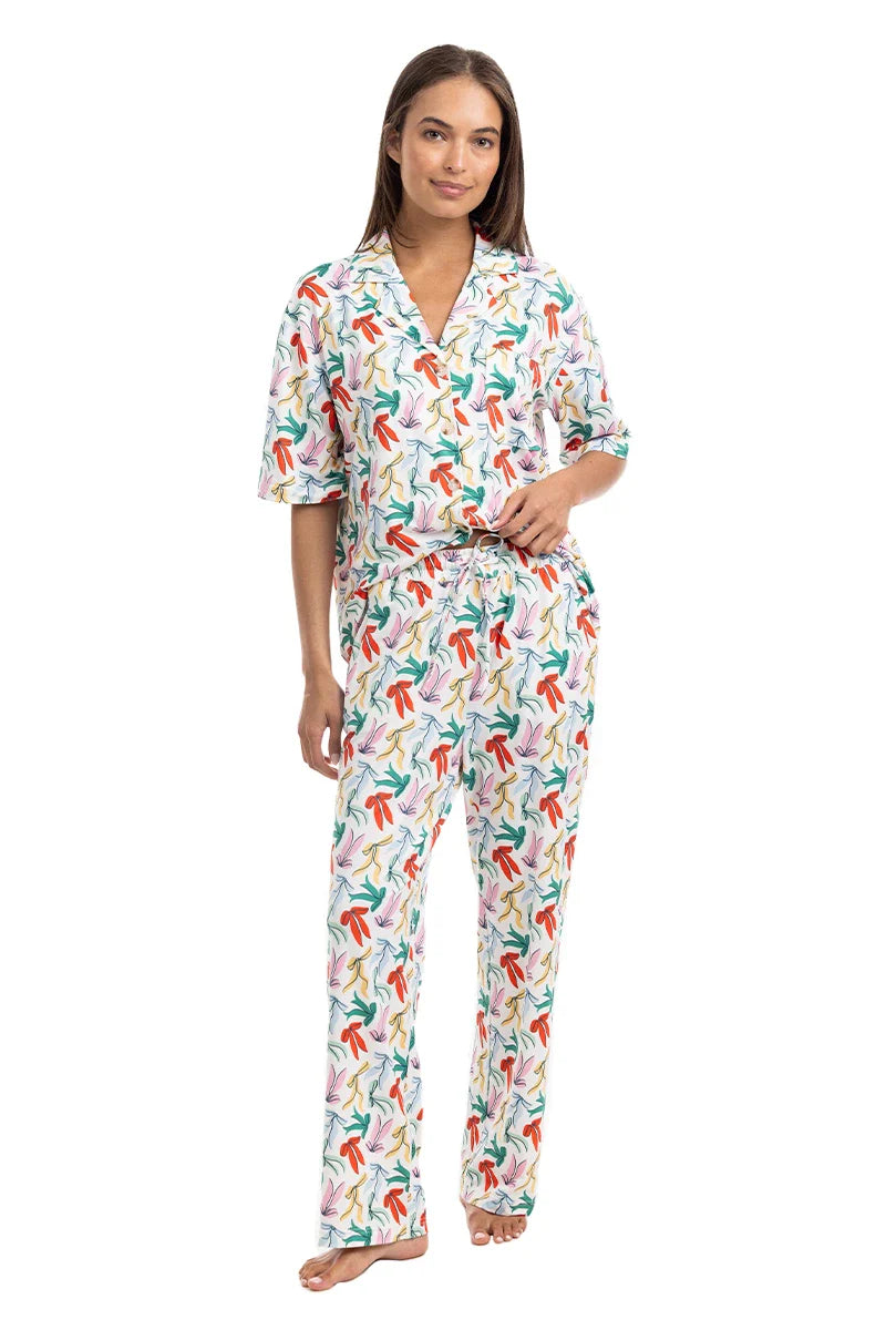 A woman stands barefoot in Katydid’s Whimsical Bow Pajamas Set, featuring a colorful abstract print with a short-sleeve button-up top and long pants—perfect festive holiday loungewear—on a white background.