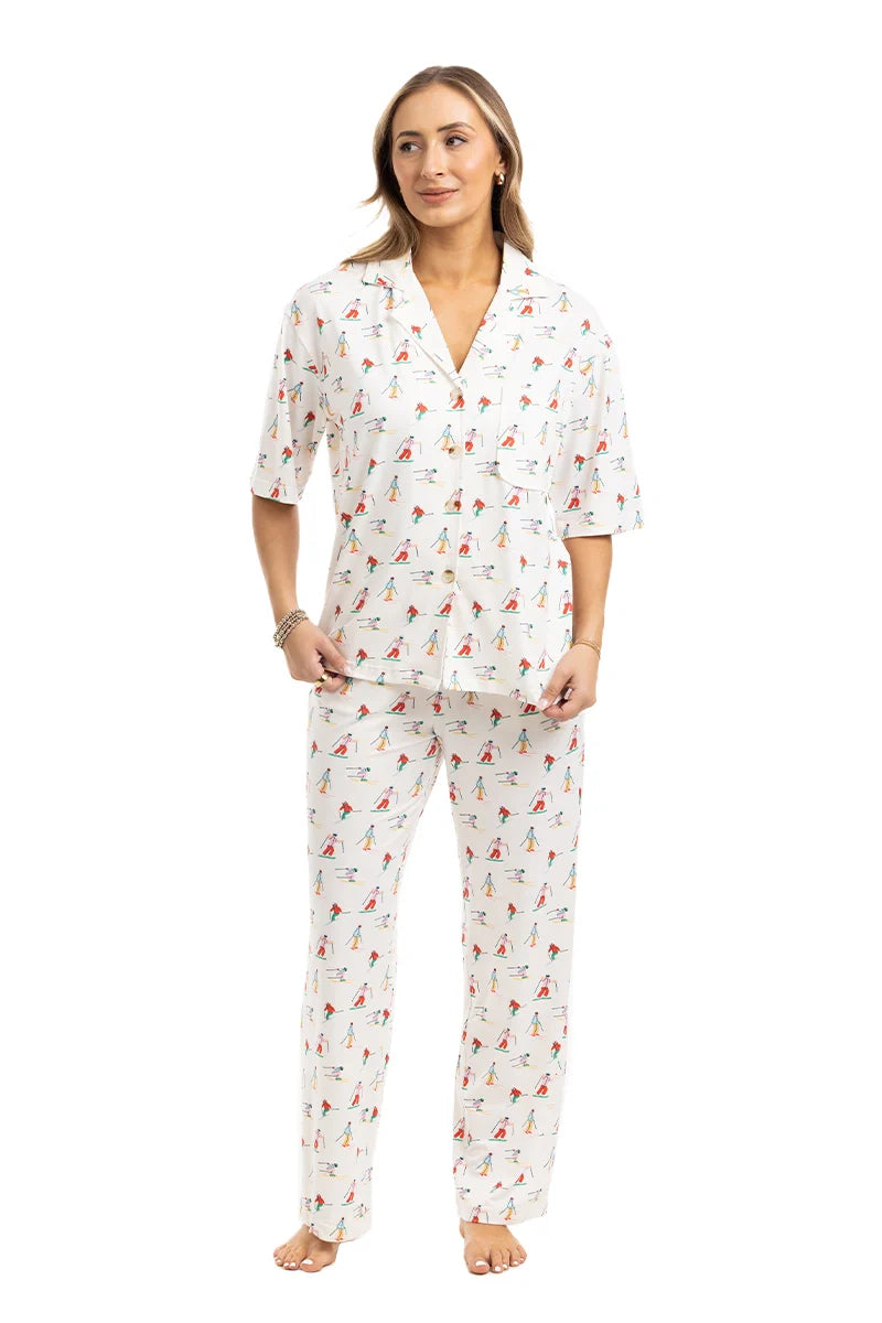 A woman with light brown hair and a relaxed expression wears the Katydid Apres Ski Matching Pajama Pants Set: a cozy, white winter set with a collared short-sleeve button-up top and long pants featuring a playful ski print. She stands barefoot.