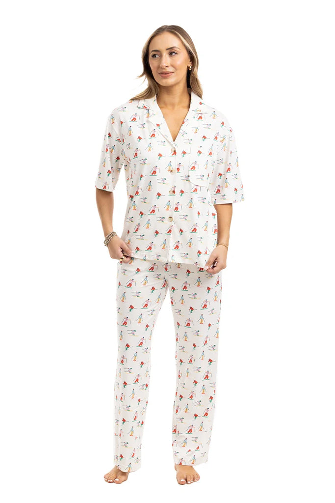 A woman with light brown hair and a relaxed expression wears the Katydid Apres Ski Matching Pajama Pants Set: a cozy, white winter set with a collared short-sleeve button-up top and long pants featuring a playful ski print. She stands barefoot.