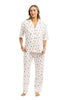 A woman with light brown hair and a relaxed expression wears the Katydid Apres Ski Matching Pajama Pants Set: a cozy, white winter set with a collared short-sleeve button-up top and long pants featuring a playful ski print. She stands barefoot.
