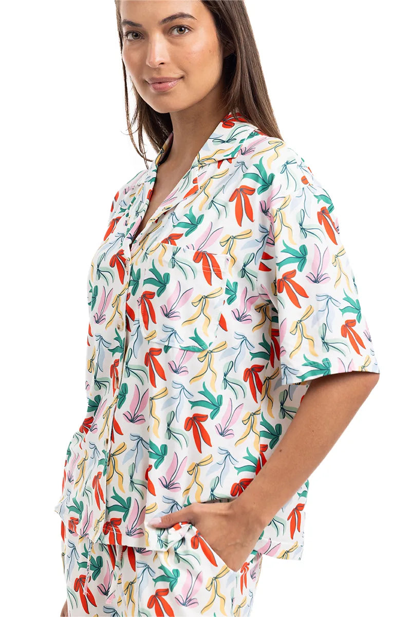 A woman wears the Katydid Whimsical Bow Pajamas Set, a colorful short-sleeved set with a playful bow print, standing with one hand in her pocket and smiling—perfect for festive holiday lounging.