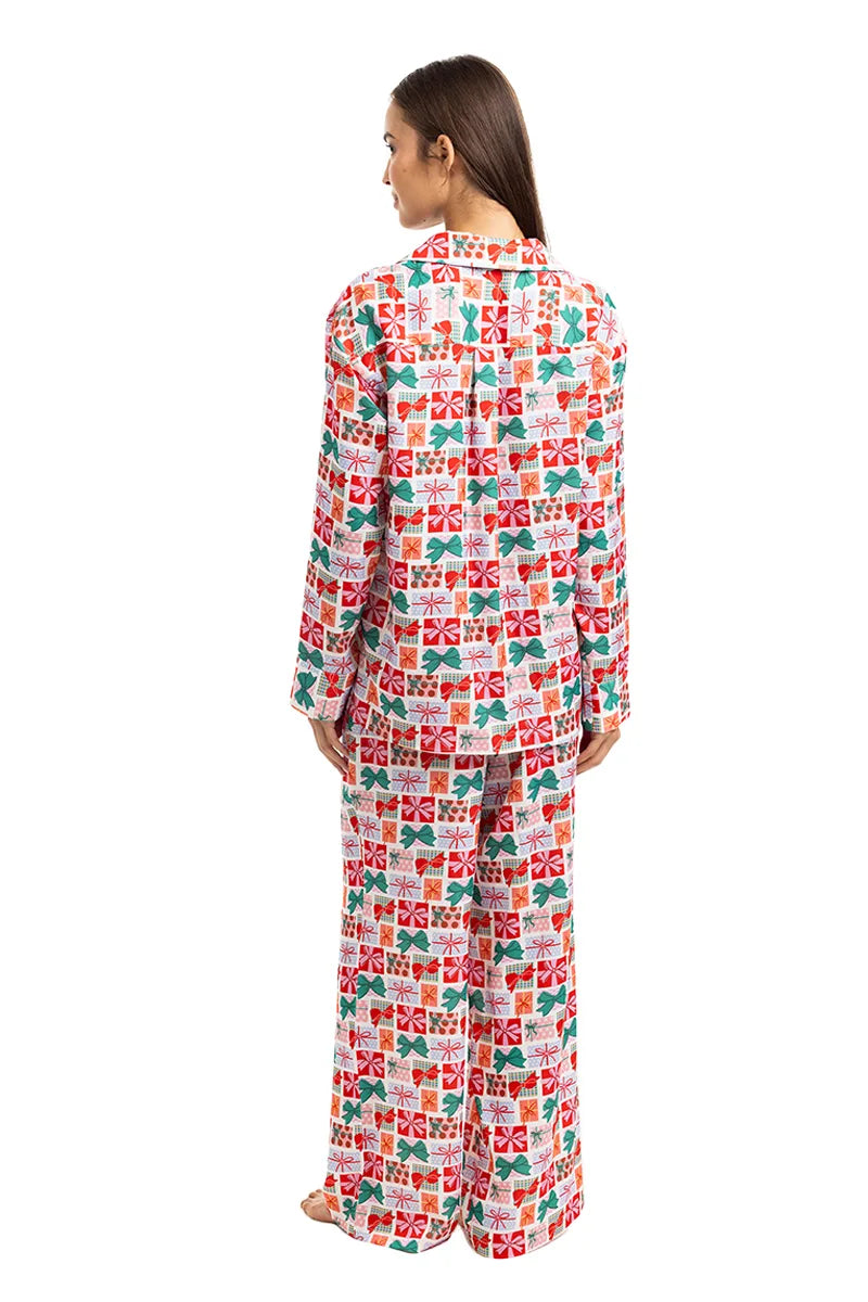 A woman stands barefoot facing away, wearing the Katydid Wrapped Presents with Bows Pajama Pants Set, which includes a matching long-sleeve top and pajama pants adorned with festive red, green, and white gift box patterns on white.