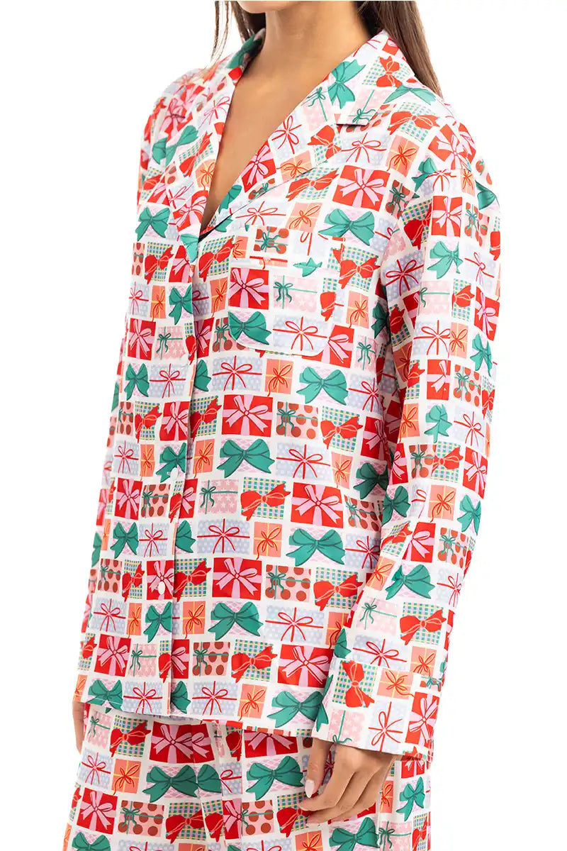 A woman wears the Katydid Wrapped Presents with Bows Pajama Pants Set, featuring festive red, green, and orange gift box and bow patterns on white. The set includes a collared button-up top and matching holiday pajama pants.