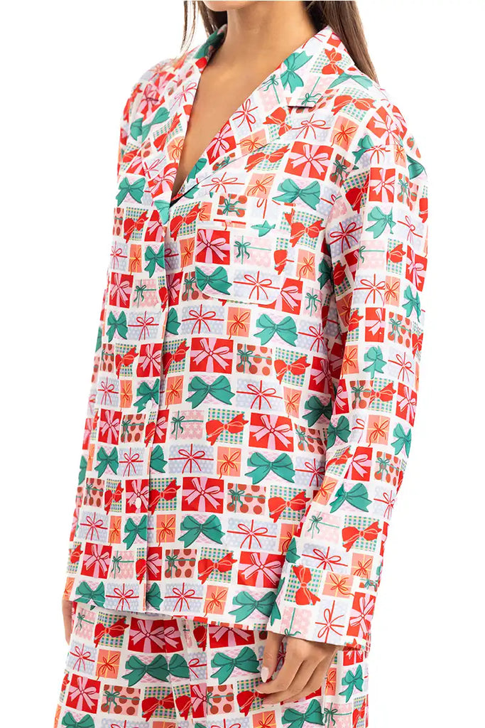 A woman wears the Katydid Wrapped Presents with Bows Pajama Pants Set, featuring festive red, green, and orange gift box and bow patterns on white. The set includes a collared button-up top and matching holiday pajama pants.