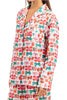 A woman wears the Katydid Wrapped Presents with Bows Pajama Pants Set, featuring festive red, green, and orange gift box and bow patterns on white. The set includes a collared button-up top and matching holiday pajama pants.