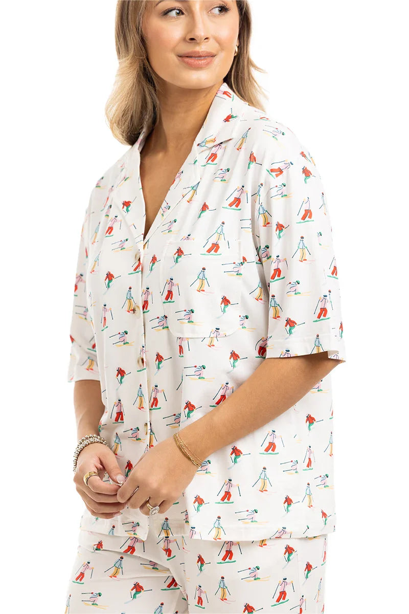 A woman wears the Katydid Apres Ski Matching Pajama Pants Set, a cream-colored set with a playful ski-inspired print of colorful skiers, featuring a notched collar and short sleeves. She stands with her hands gently clasped.