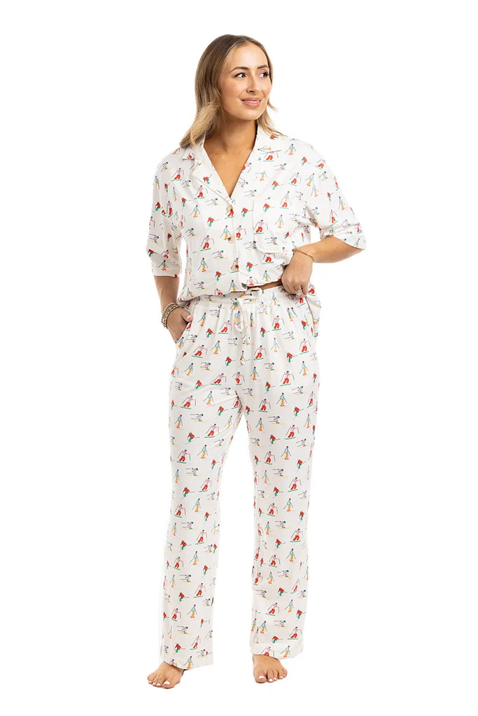 A woman stands barefoot wearing the Katydid Apres Ski Matching Pajama Pants Set—a white pajama set with a colorful ski print, featuring a short-sleeve button-up shirt and long elastic-waist pants. She smiles and looks slightly to the side.