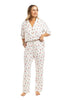 A woman stands barefoot wearing the Katydid Apres Ski Matching Pajama Pants Set—a white pajama set with a colorful ski print, featuring a short-sleeve button-up shirt and long elastic-waist pants. She smiles and looks slightly to the side.