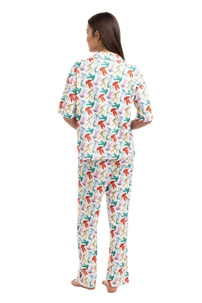 A woman stands barefoot facing away, wearing the Katydid Whimsical Bow Pajamas Set featuring a colorful abstract pattern of red, green, blue, and orange shapes on a white background.