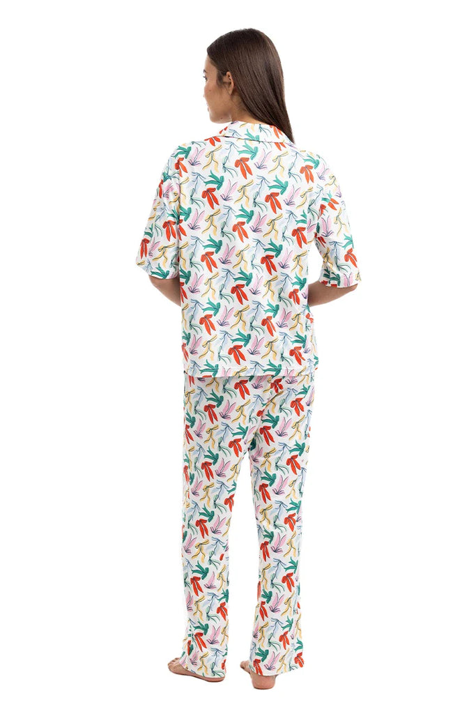 A woman stands barefoot facing away, wearing the Katydid Whimsical Bow Pajamas Set featuring a colorful abstract pattern of red, green, blue, and orange shapes on a white background.
