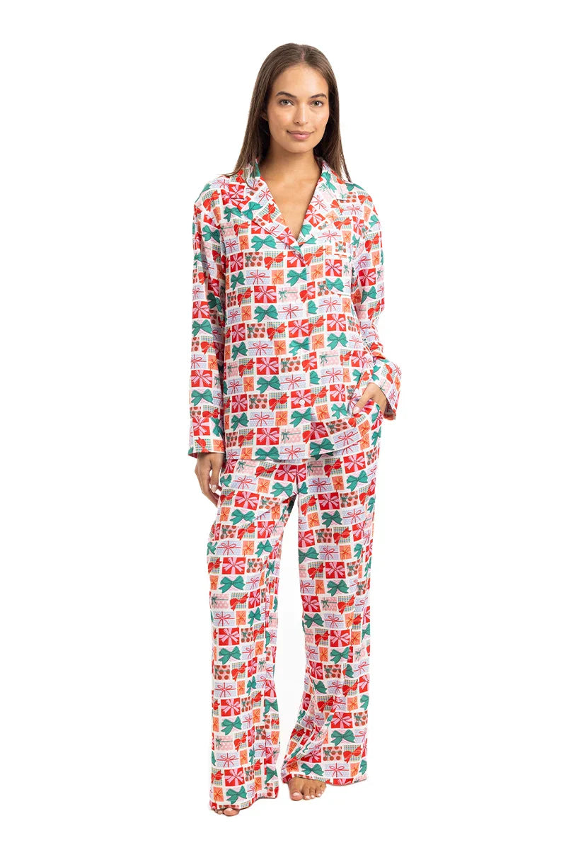 A woman smiles with her hand in one pocket, wearing the Katydid Wrapped Presents with Bows Pajama Pants Set—a festive, long-sleeve button-up top and loose pajama pants featuring a colorful gift box pattern.